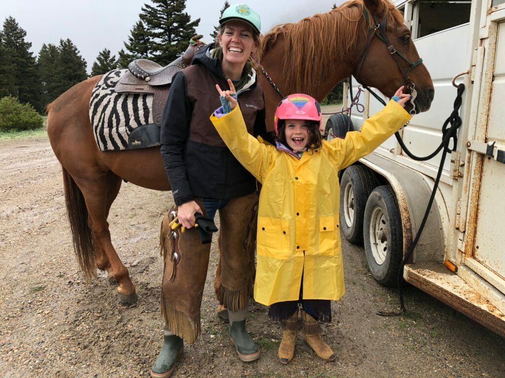 A little girl wearing a yellow rain slicker stands with her mom and a horse on a cloudy Montana day.
