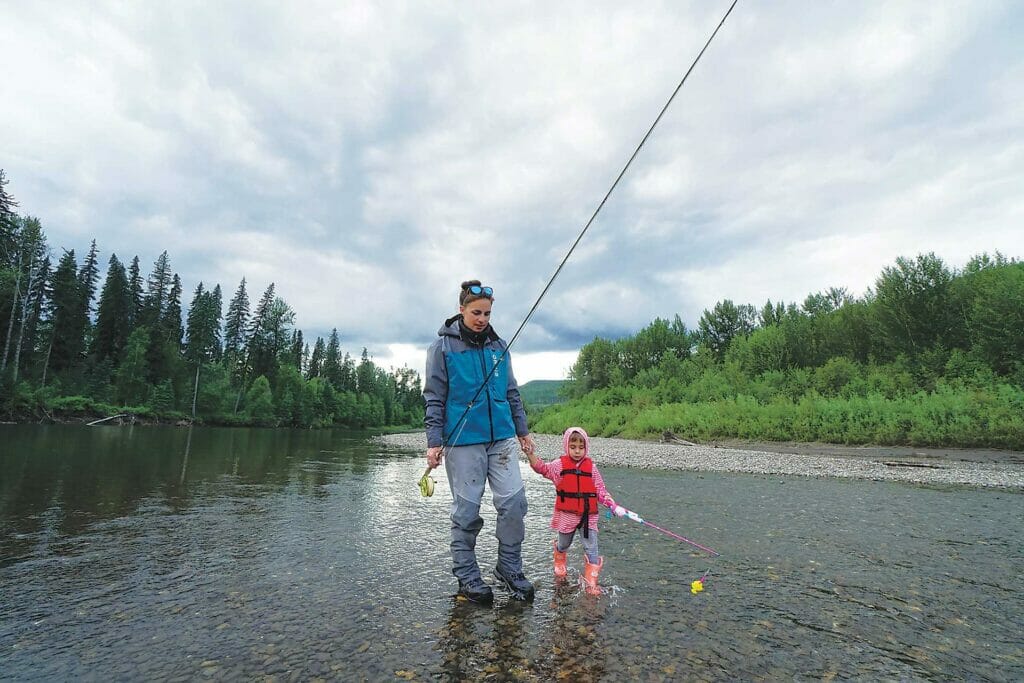 A mother and daughter wade in a trout stream carrying fishing rods.