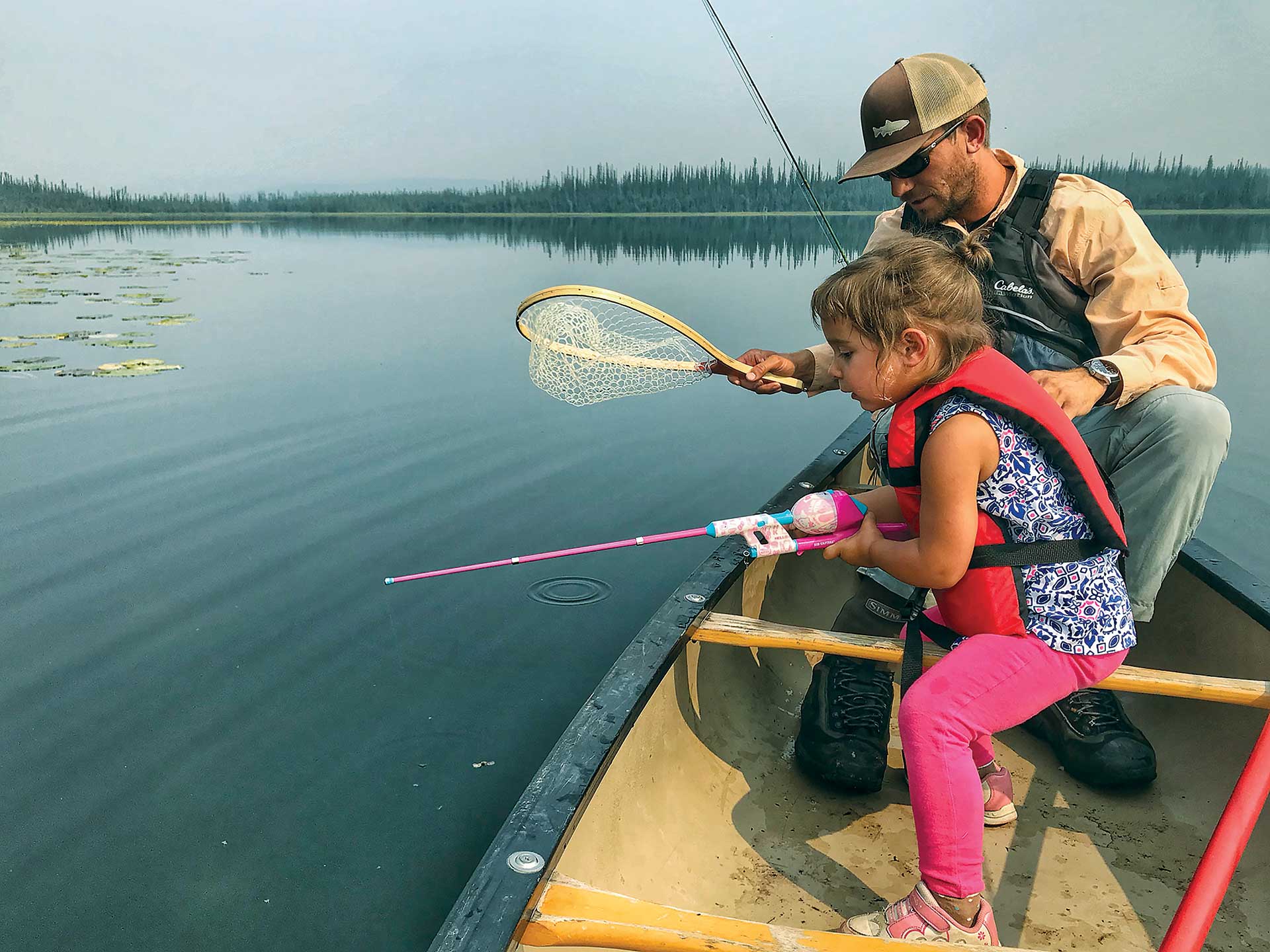 A young girl in a canoe works to land a trout with her father.