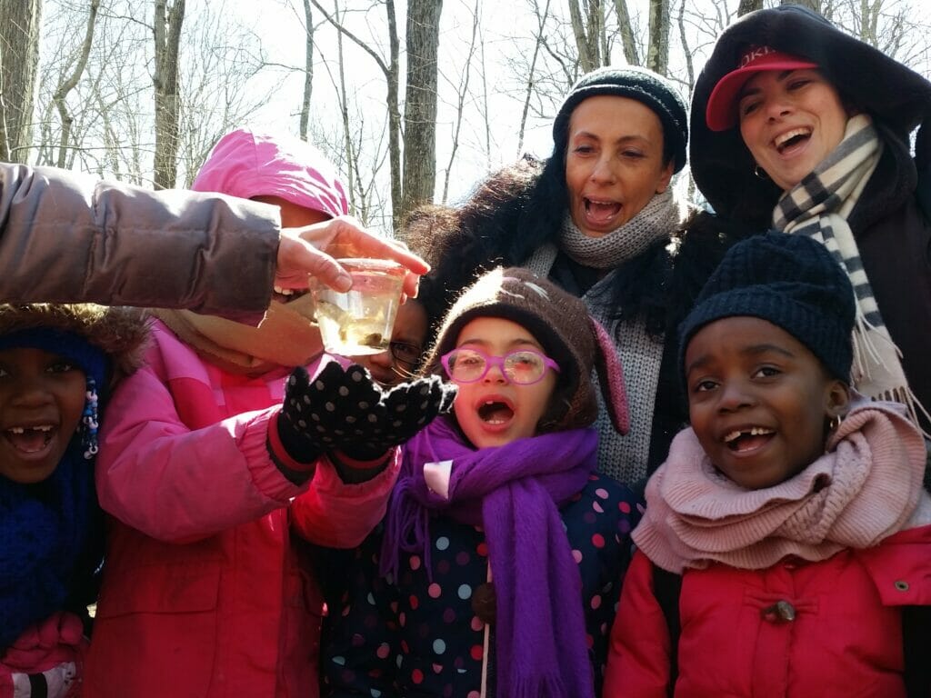 Group of elementary kids and adults smiling at small trout in cup of water