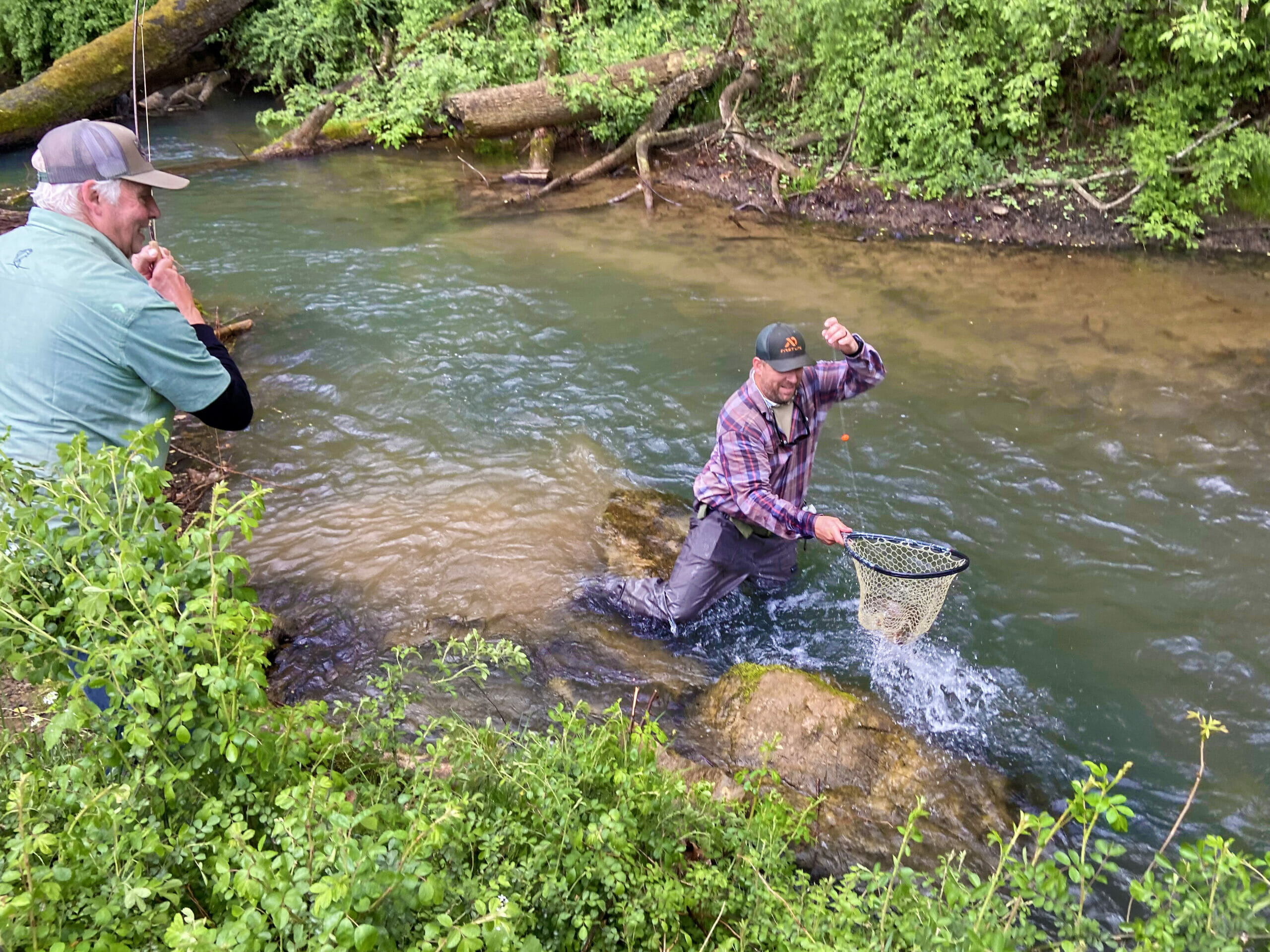 Man in river gets fish into net while his old friend watches