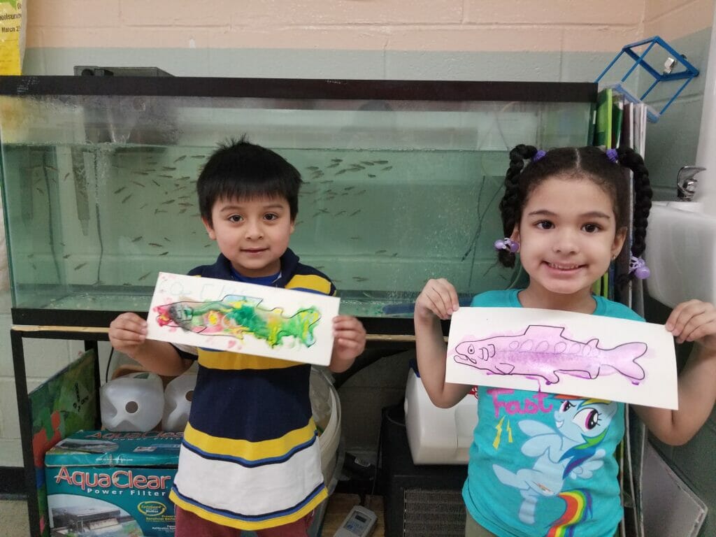 Two elementary students hold drawings of fish in front of classroom fish tank.