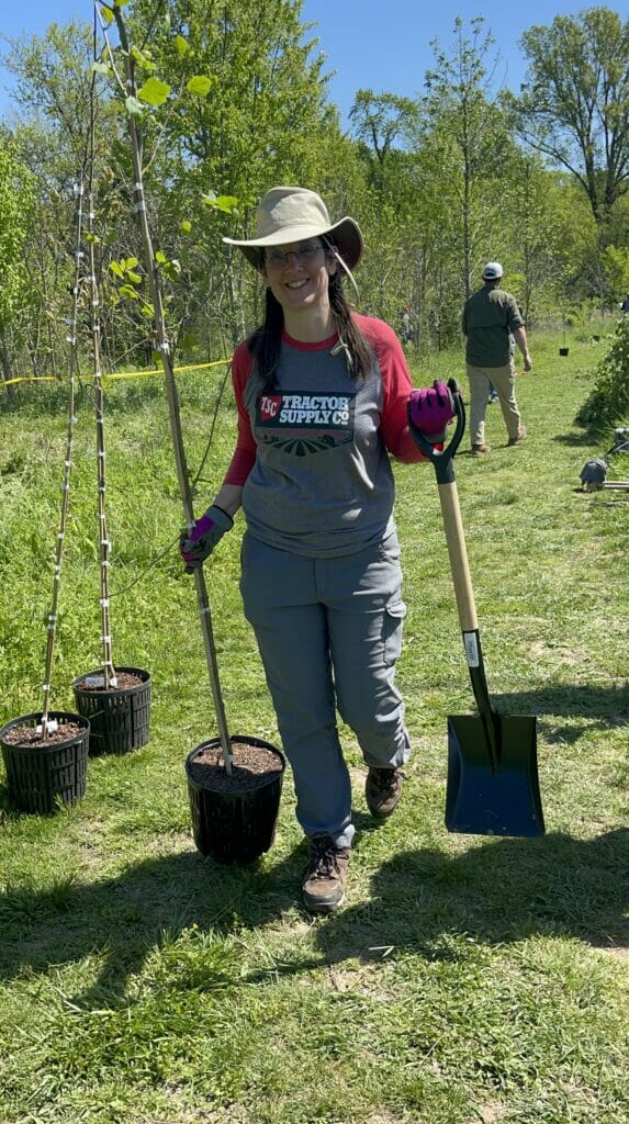 A smiling woman carries a potted tree and shovel