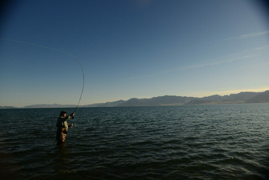 Man casts while standing in beautiful lake with mountains in the background