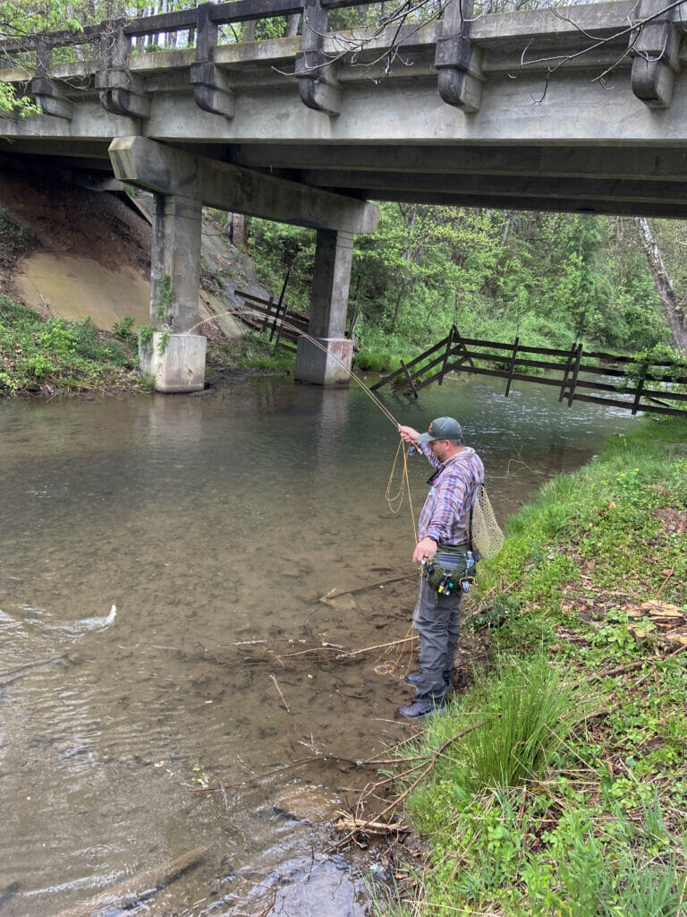 Man stands next to creek and bridge pulling in a fish