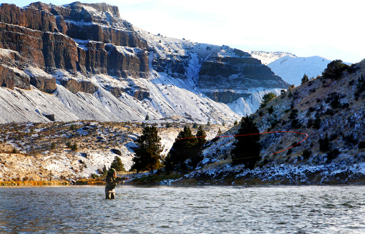 Person in wide river casting with snowy mountains behind