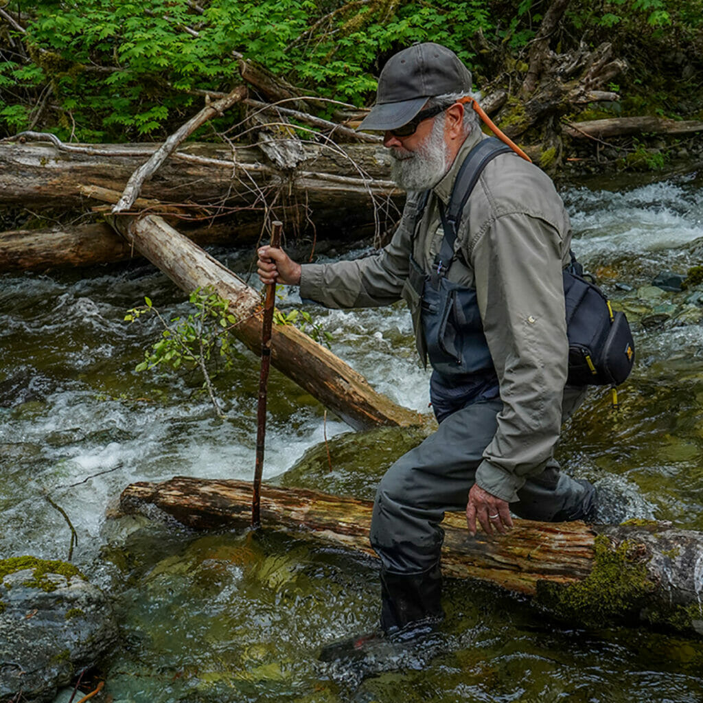 Man with beard and walking stick walks through rushing water