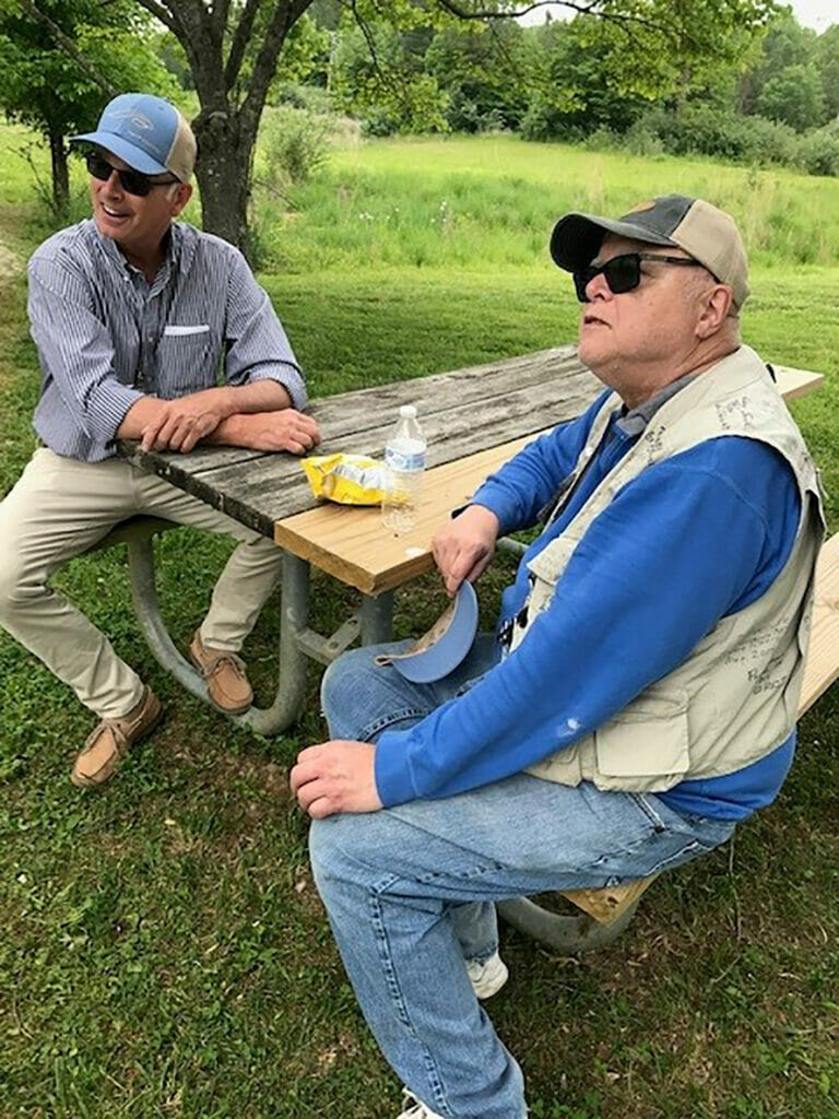 Two men sit at a picnic table with a bottle of water and bag of chips