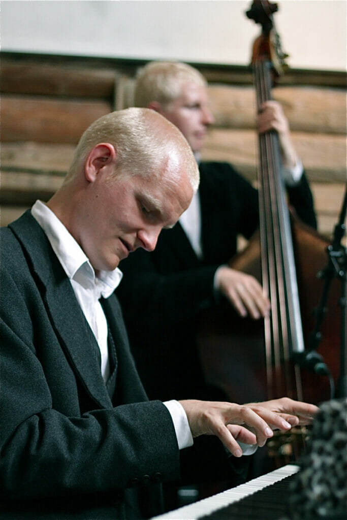 Man in suit plays piano while his brother plays base in the background