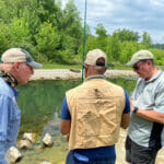 Three men stand by a river looking at the same thing