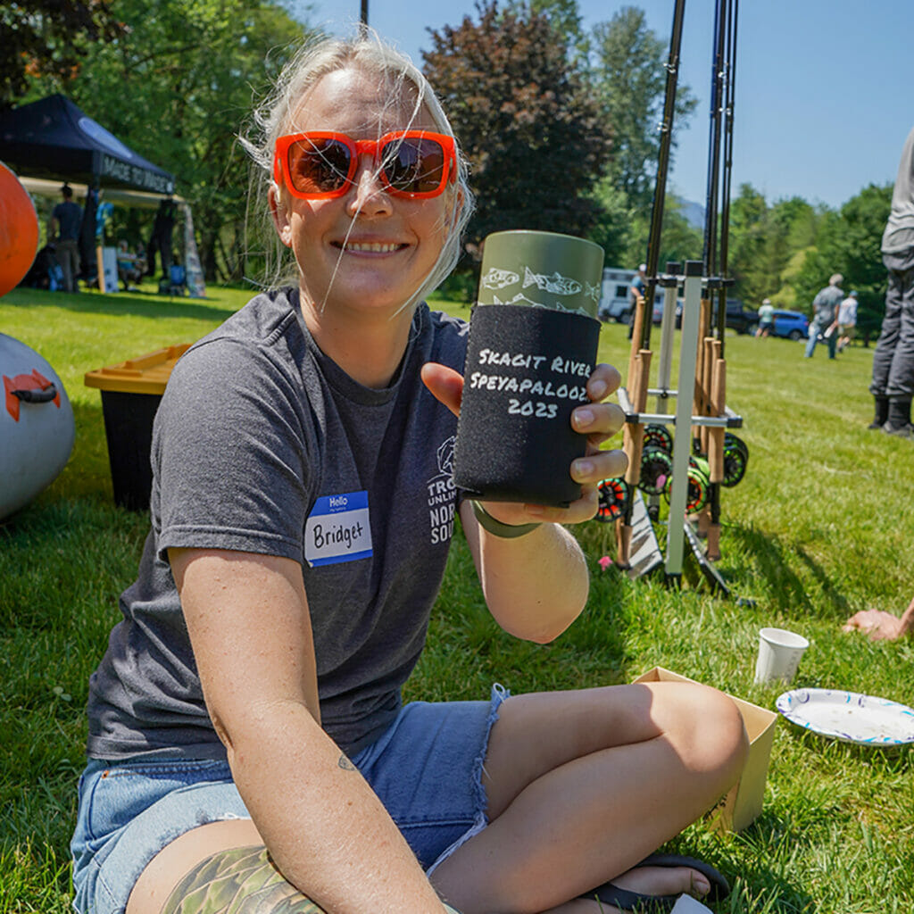 Woman with orange sunglasses shows off koozie reading: Skarit River Speyapalooza 2023