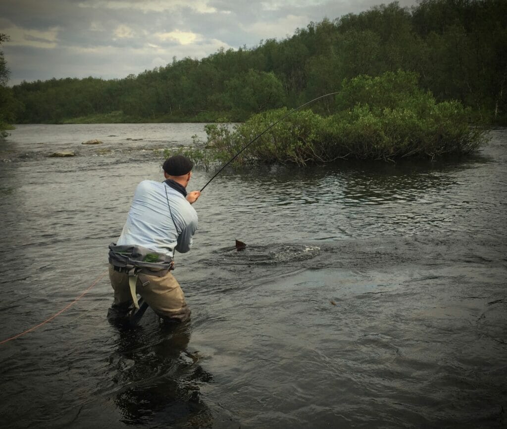 Man in stream with a cool hat reeling in a fish