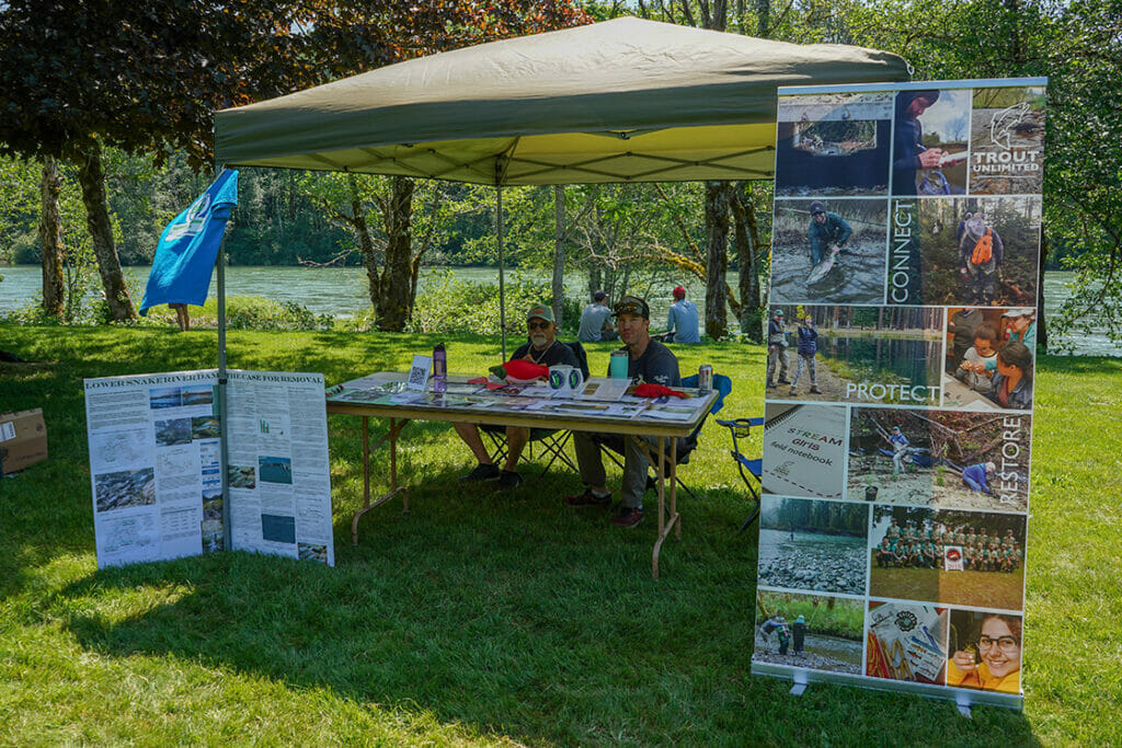 Two men sit at a TU volunteer booth next to a river