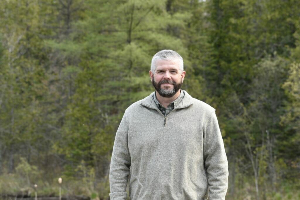 Man in fleece jacket smiles at camera in front of some trees