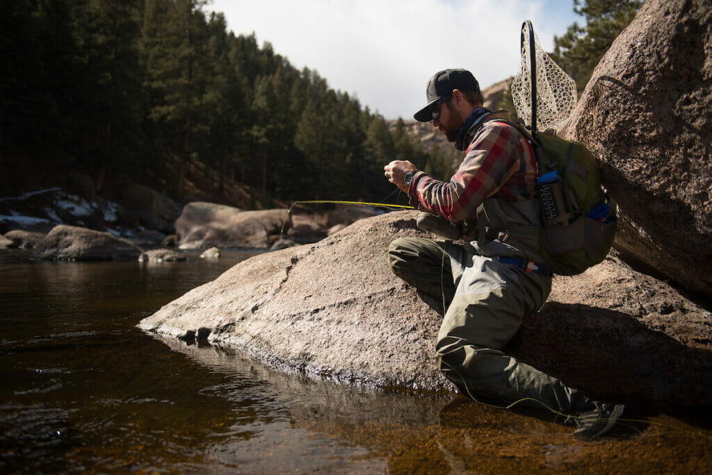 Man kneels next to stream changing his fly