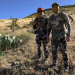 Two men wearing camoflauge smile on a mountain hill.