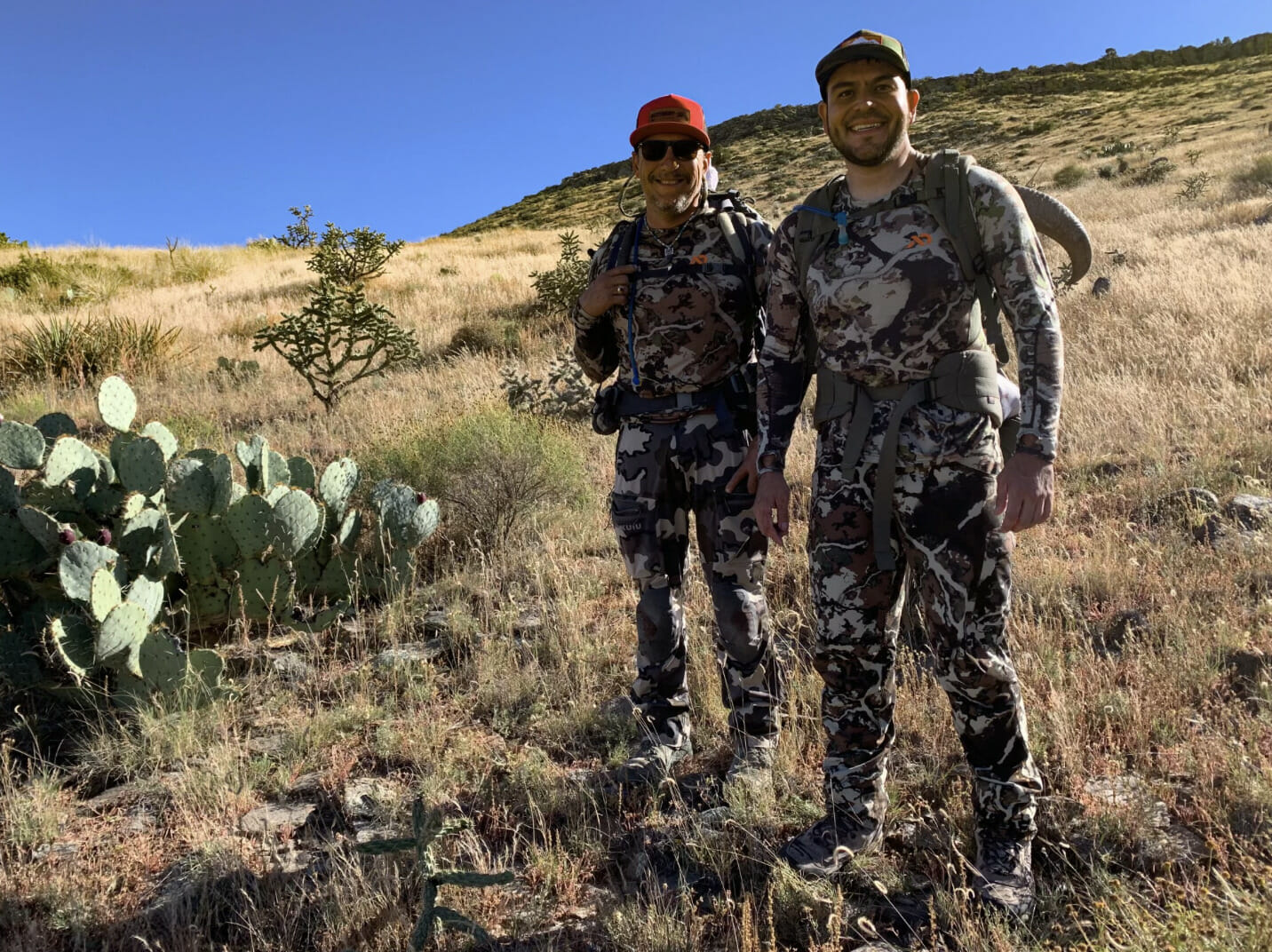 Two men wearing camoflauge smile on a mountain hill.