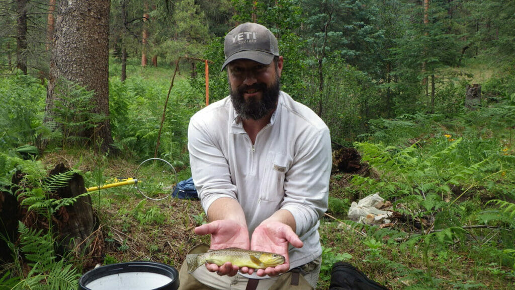 A smiling man holds a dead fish in his hands in the woods.