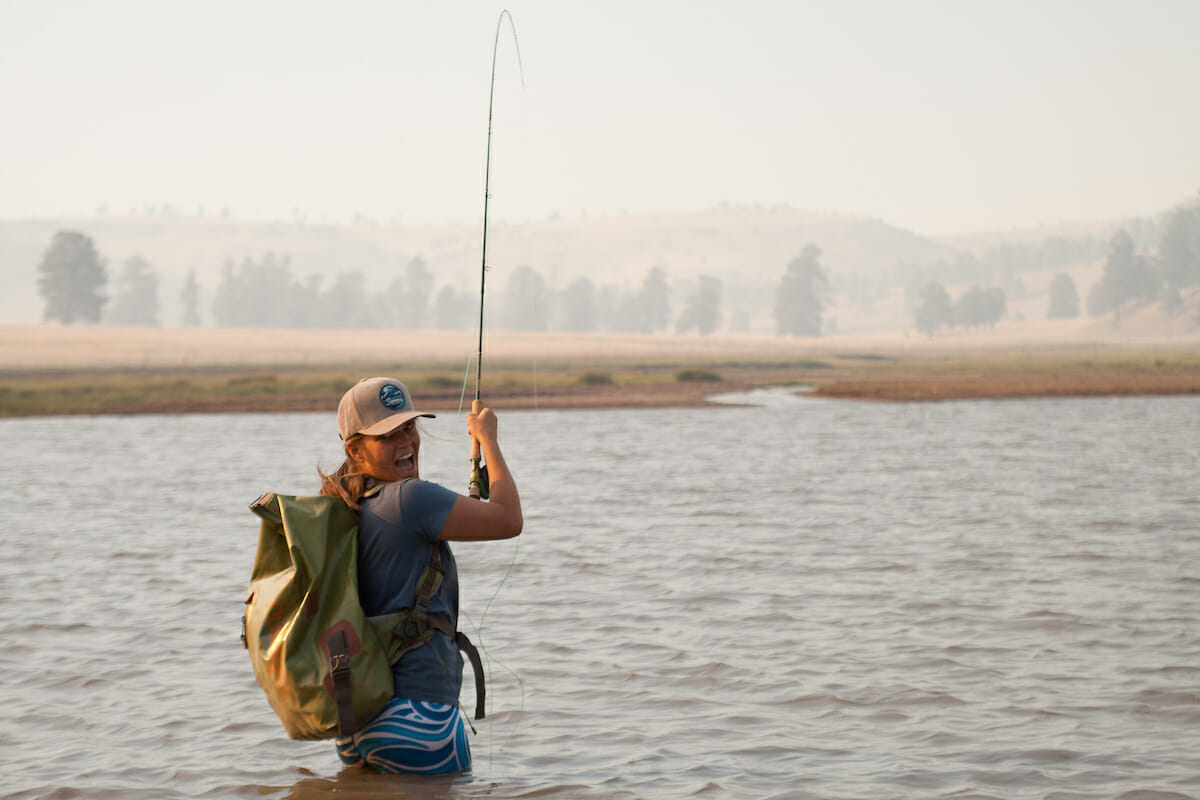 Woman in a river smiling at camera as she has a fish on her line