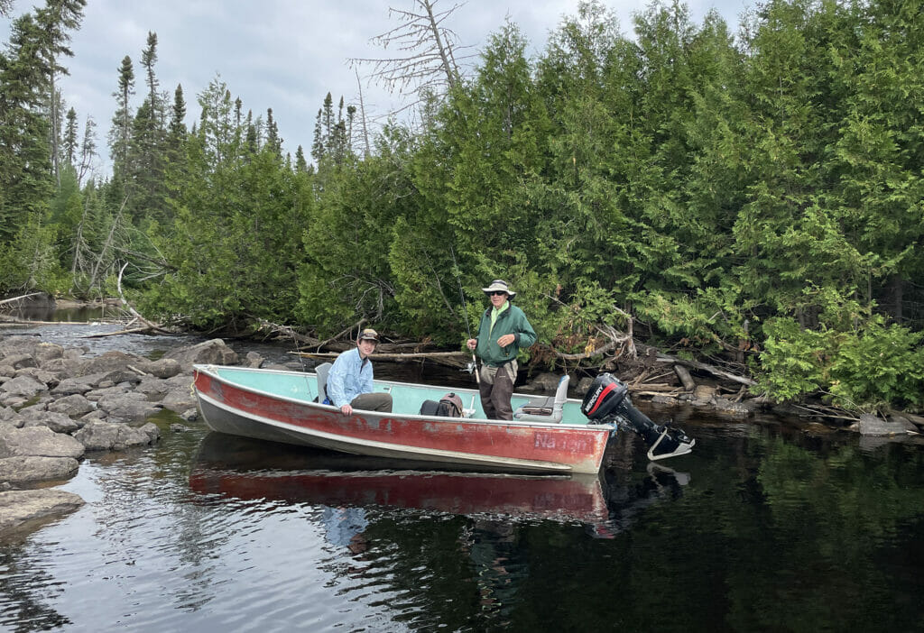 An old man stands up in a beat-up motor boat with a young man sitting