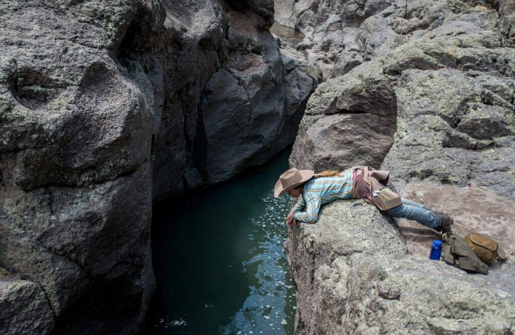 A woman with a cowboy had leans over a rocky wall looking at the river