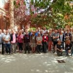 A group of around two dozen people stand on a shady sidewalk
