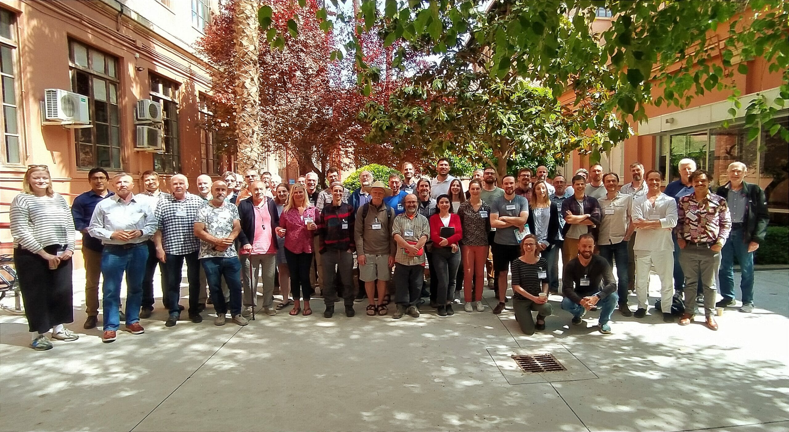 A group of around two dozen people stand on a shady sidewalk