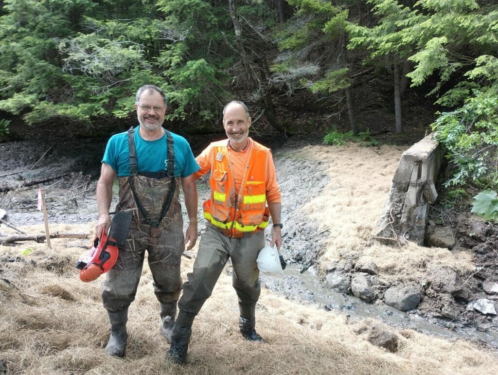 Two smiling men stand by a muddy creek