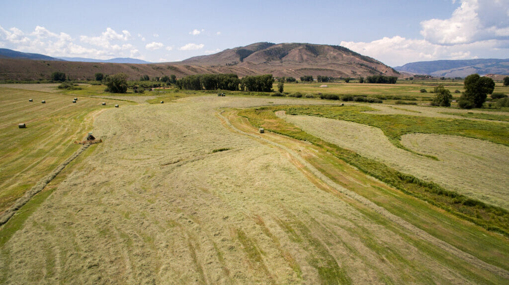 A wide view of a tractor turning cut hay into round bales with mountains in the background and they all need water