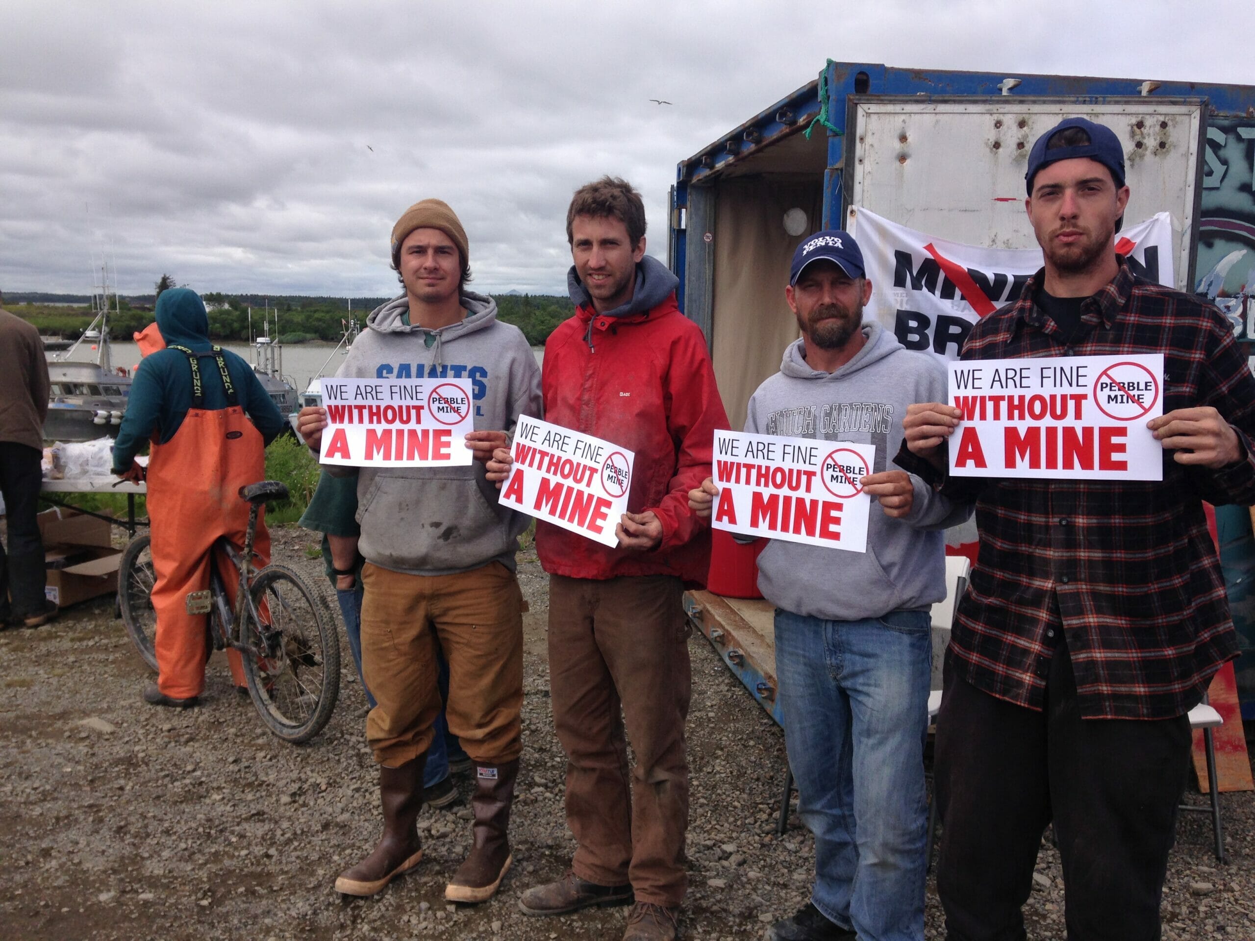 Four men stand with signs reading "We are fine without a mine. No Pebble Mine"