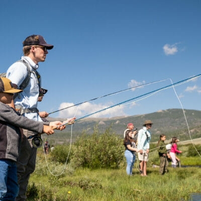 Several college students fish on a river bank with several younger kids