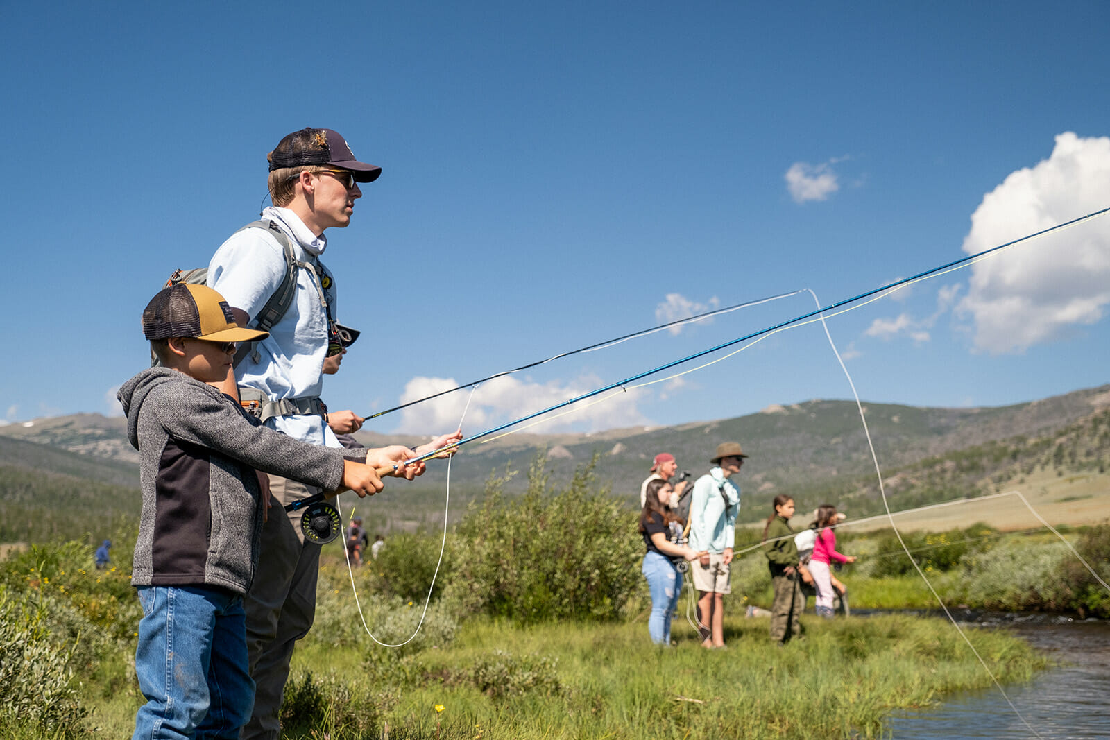 Several college students fish on a river bank with several younger kids