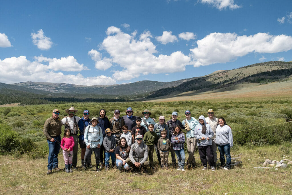 A group of people in an Ambassador Summit, including children, stand in front of moutains