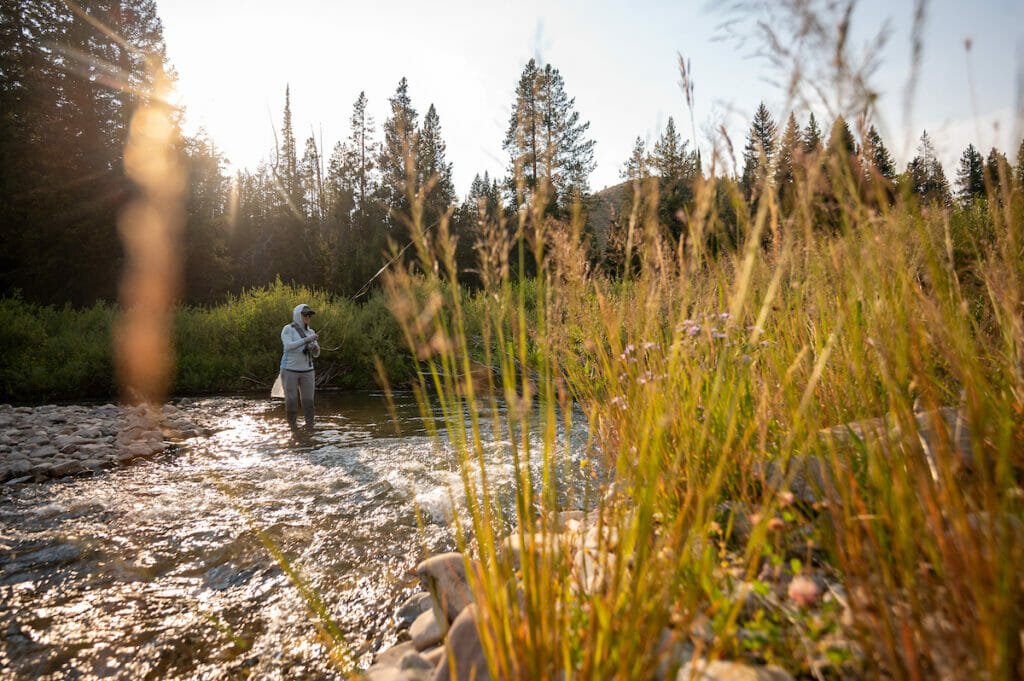 Woman stands in a rushing river fishing with the setting sun behind her working on casting accuracy