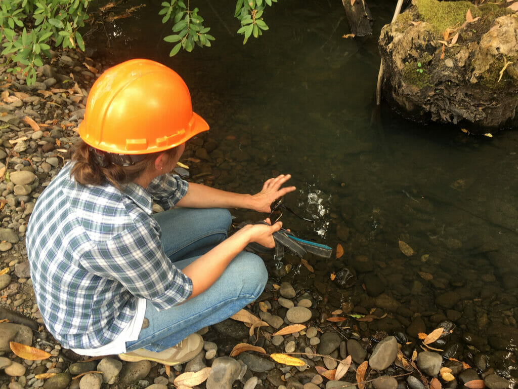 Woman with hard hat crouches next to stream