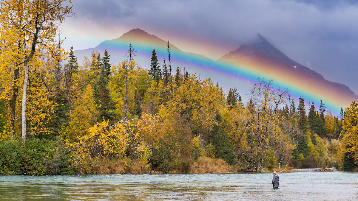 Distant view of a man fishing in a deep river with a rainbow and mountain in the background.