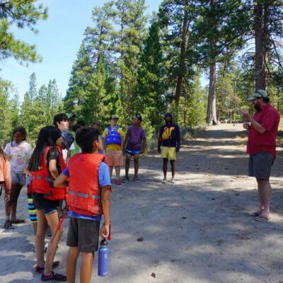 Man talks to a group of kids wearing life preservers by a river