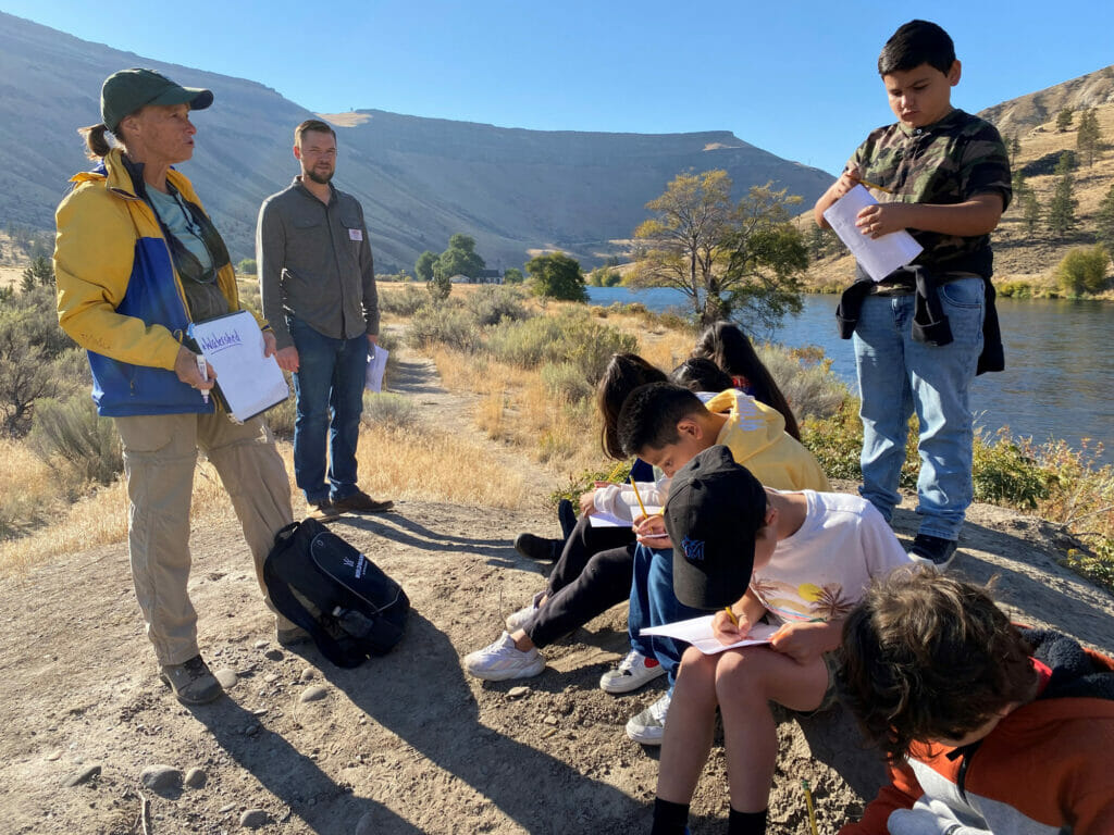 A group of kids write in notebooks next to a river while their teachers instructs education