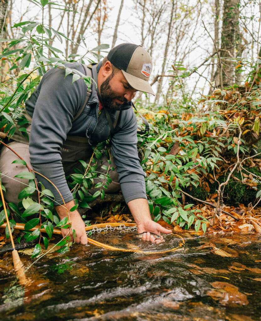 Man crouches next to stream with a fish in his hand