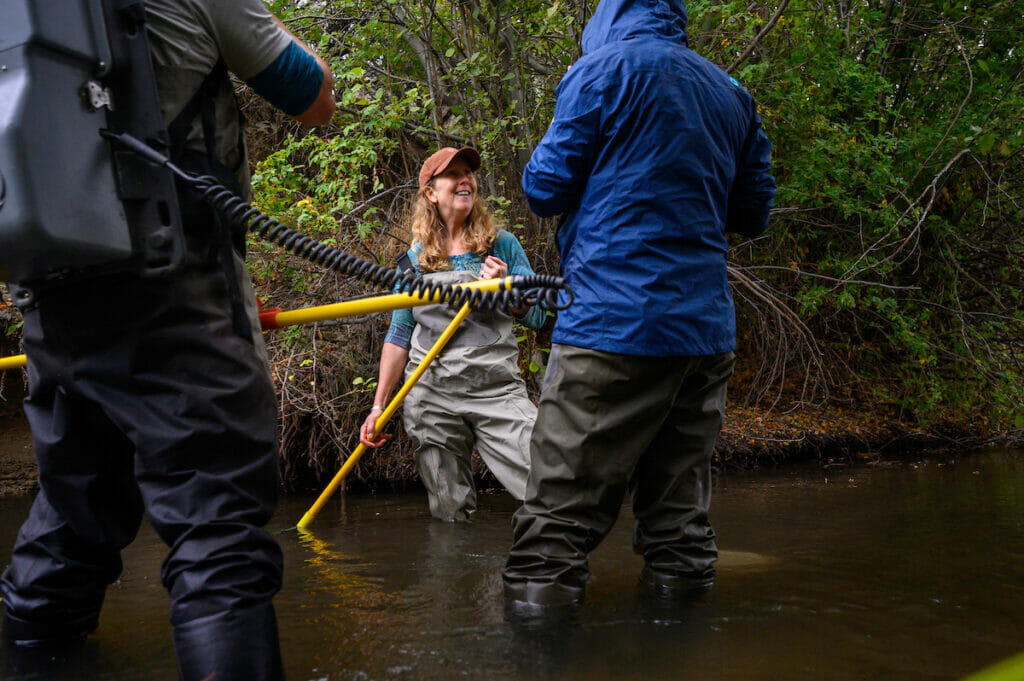 Three people wearing waders and with weird equipment stand in a stream.