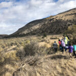A teacher and seven 3rd graders walk through a dry landscape next to a mountain