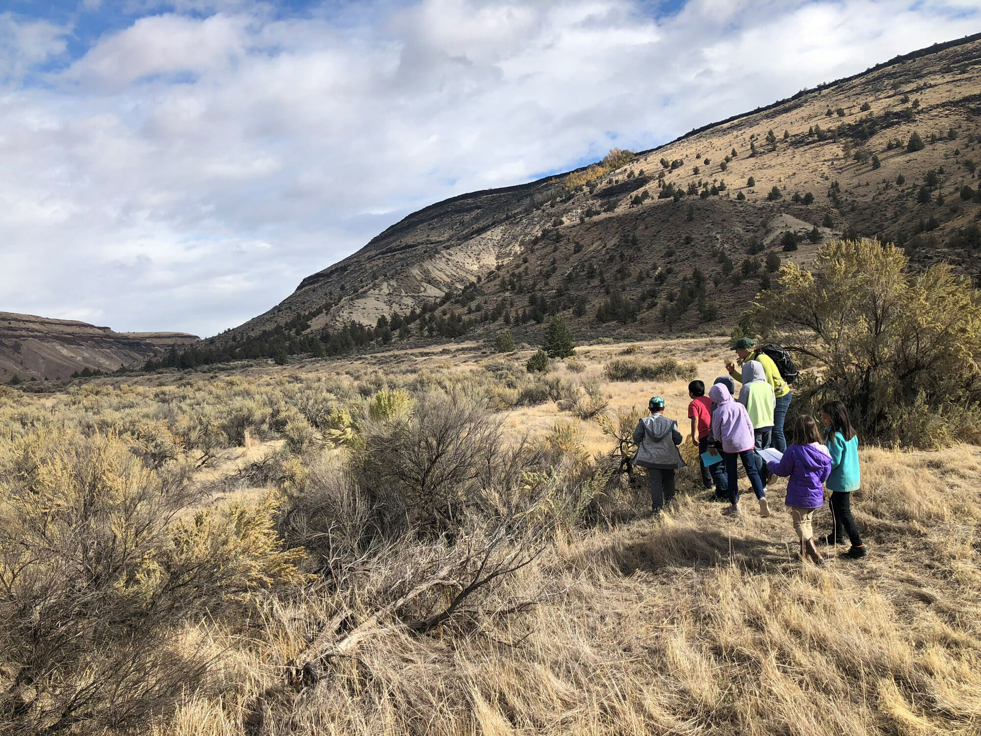 A teacher and seven 3rd graders walk through a dry landscape next to a mountain