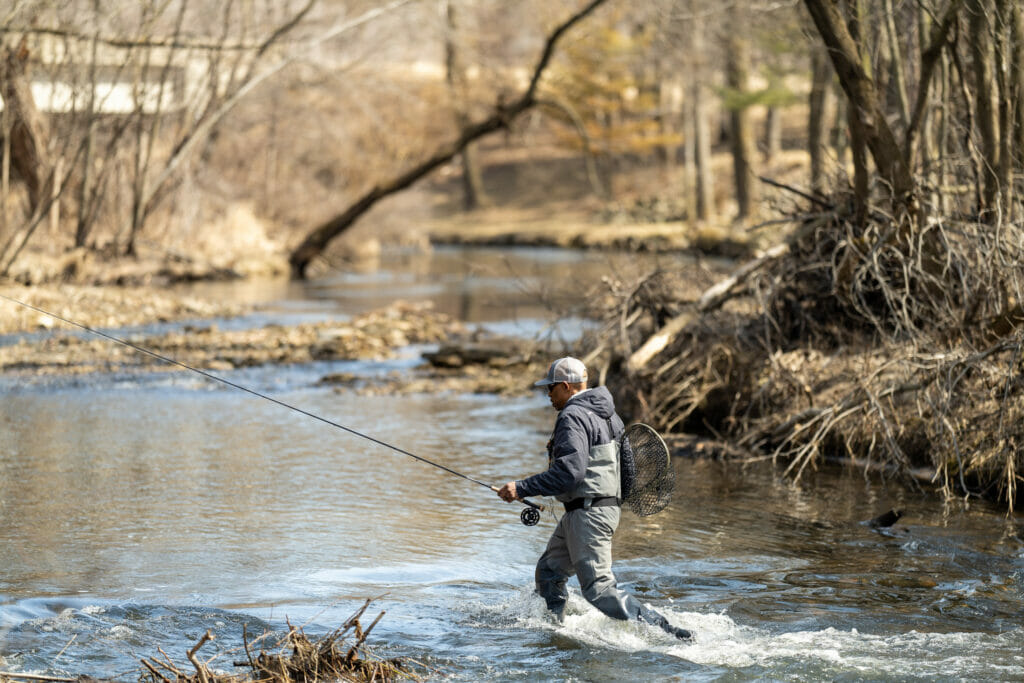 Man with waders and fishing gear walks through a stream