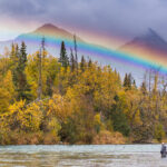 angler in river fly fishing with rainbow overhead mountains in background
