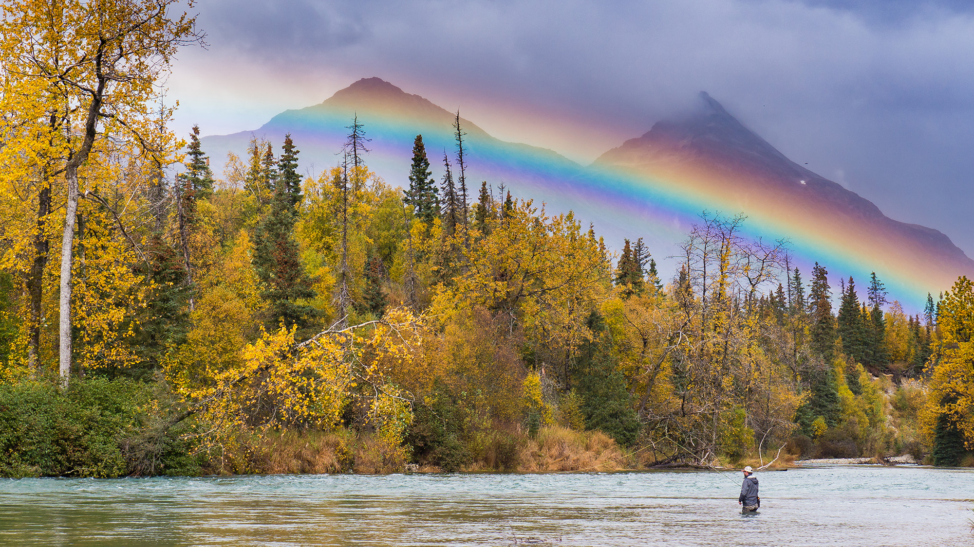 angler in river fly fishing with rainbow overhead mountains in background